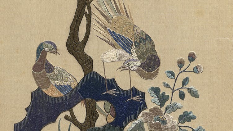 embroidery of two colorful birds on a branch. the bird on the right is bent over, looking backwards through his long legs at the bird on the left 